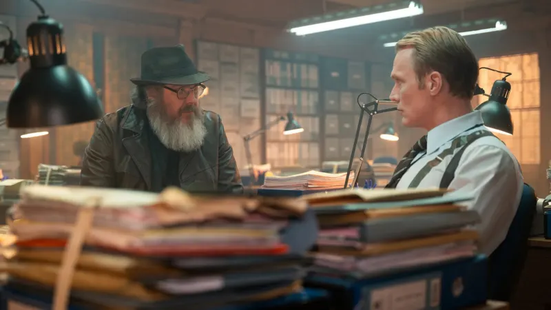 Paul Bettany jako Vision siedzi przy biurku zawalonym teczkami i rozmawia z brodatym mężczyzną w kapeluszu, kadr z serialu VisionQuest